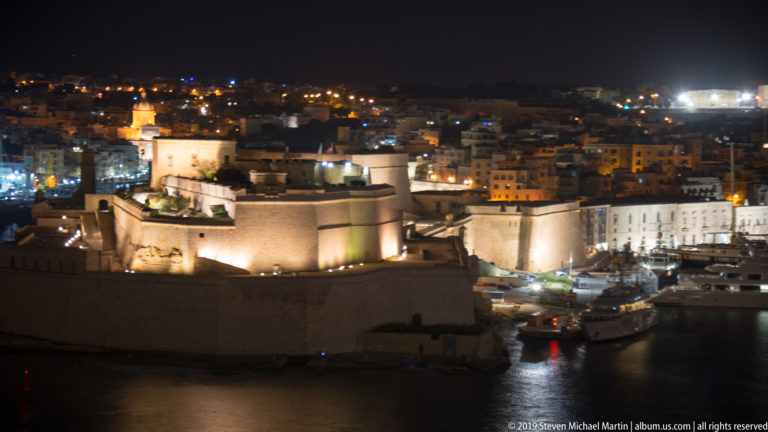 Large illuminated stone fortification on the harbor edge, with rounded defensive walls and a dense cityscape behind it at night.