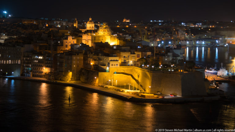 Illuminated fortified peninsula at night with dense stone buildings and church architecture reflected in dark harbor water.