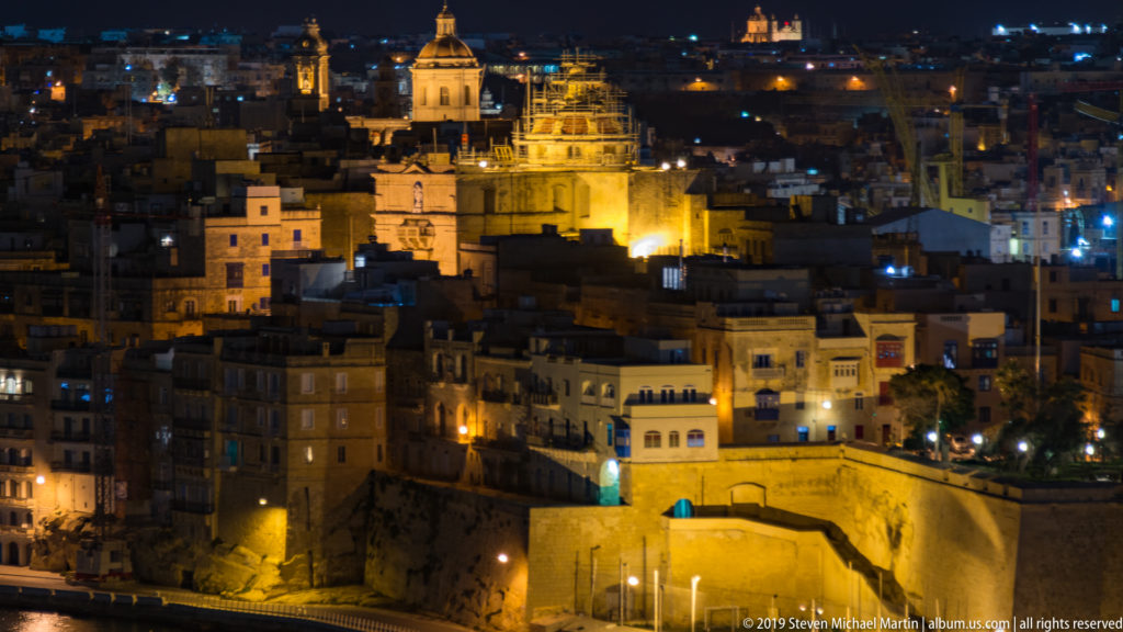 Nighttime harbor cityscape with illuminated church domes, dense stone buildings, and massive waterfront fortifications across the water.