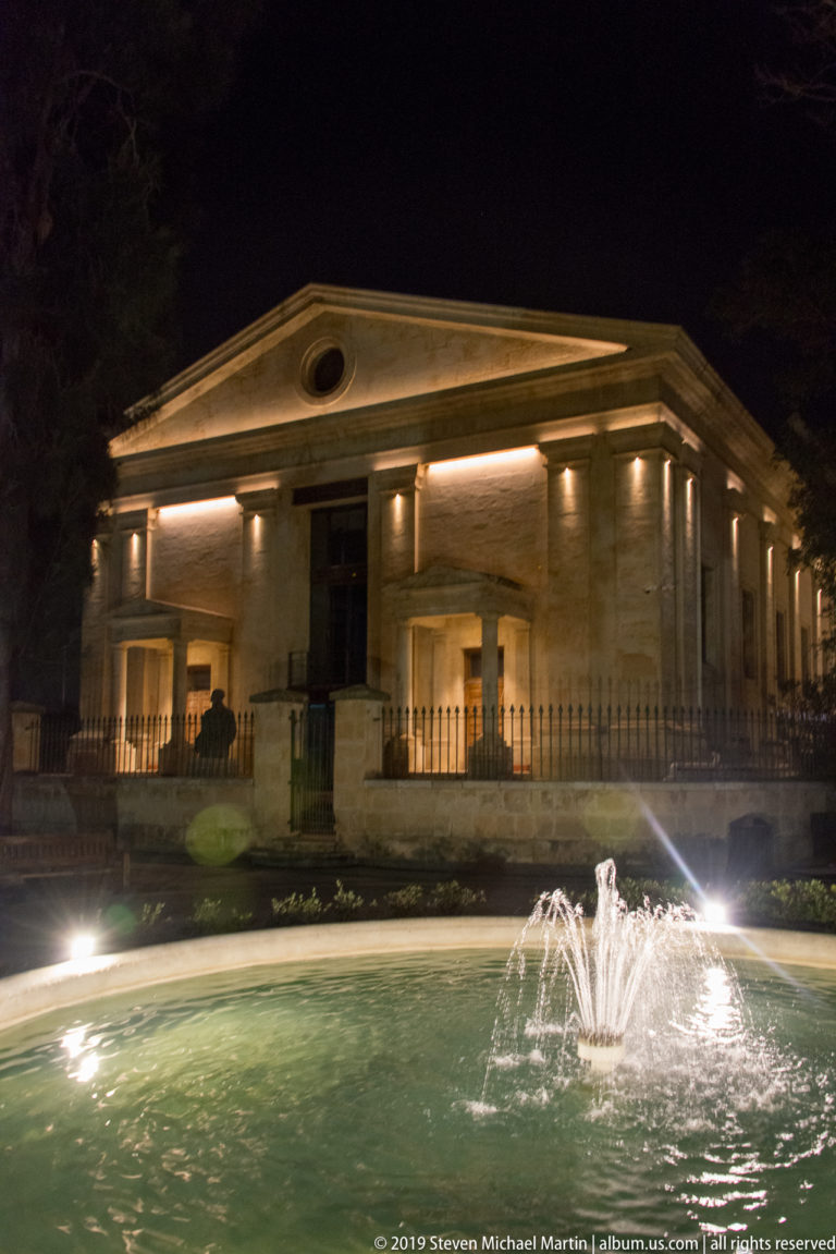 Night view of a low neoclassical stone pavilion with triangular pediment and columns behind a lit circular fountain.