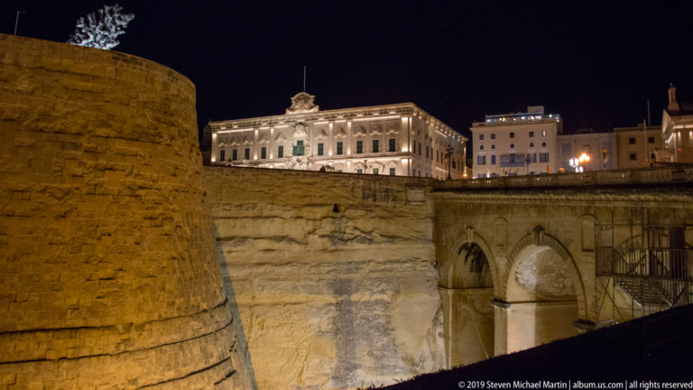 Illuminated limestone bastion and arched masonry rising from a deep rock-cut ditch, with city buildings above at night.