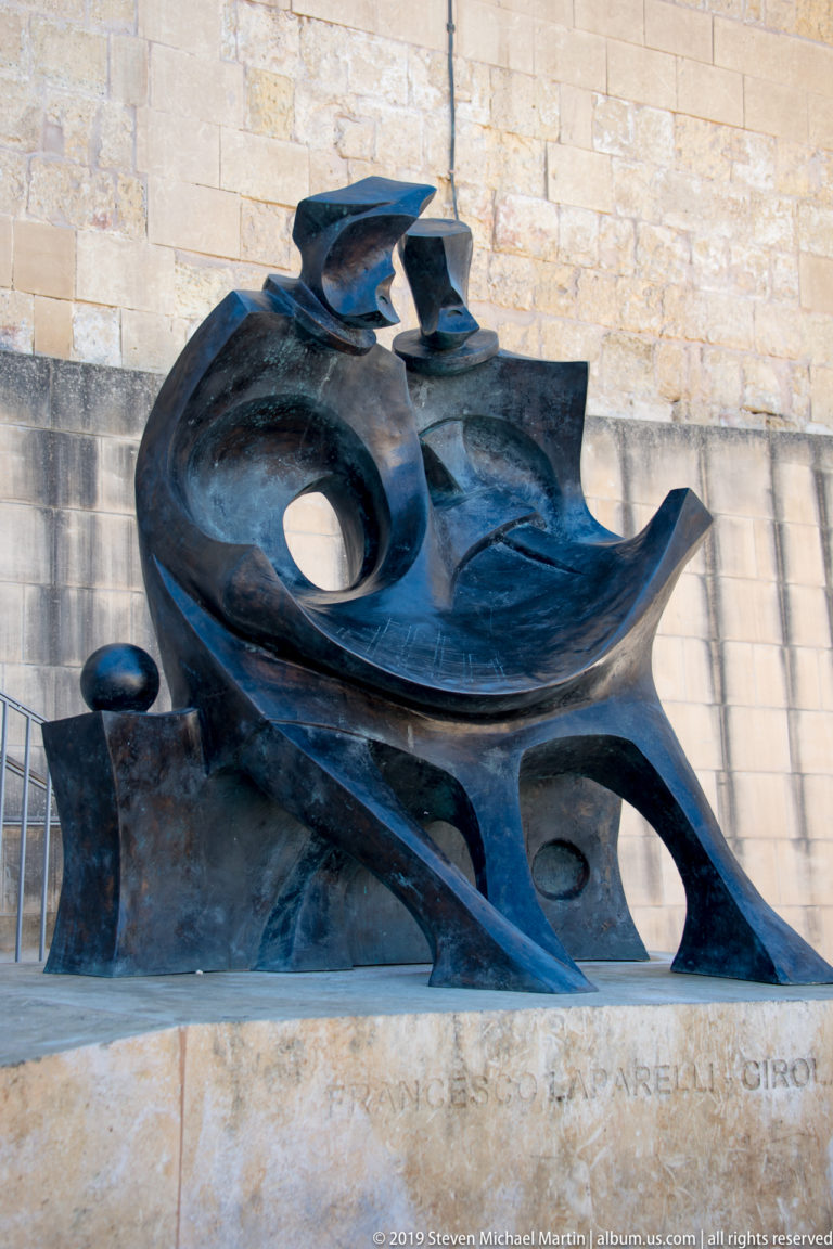 Abstract dark bronze sculpture of interlocking curved forms and circular openings on a pale stone base inscribed for Ġorġ Borg Olivier.