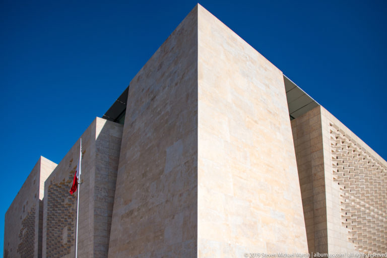 Low-angle view of contemporary pale-limestone civic building with massive geometric walls, recessed openings, perforated stone screen, and flagpoles.