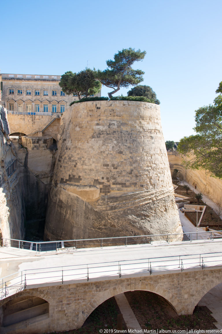 Rounded limestone bastion above a deep defensive cut, with modern railings and paths below, a windswept tree above, and buildings beyond.