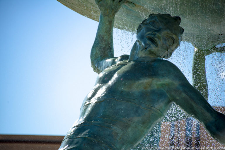 Extreme low-angle close view of a bronze Triton supporting the fountain basin, with water streaming behind his shoulder.