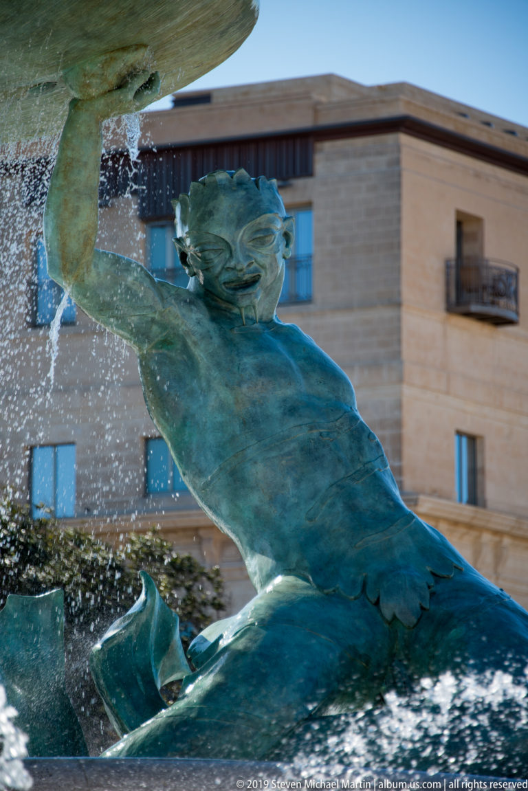 Close profile of a patinated bronze Triton raising one arm beneath a fountain basin, with water droplets and a stone building behind.