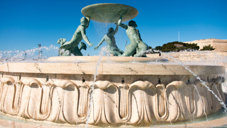 Three bronze Triton figures supporting a broad basin above a carved stone fountain base with cascading and arcing water.