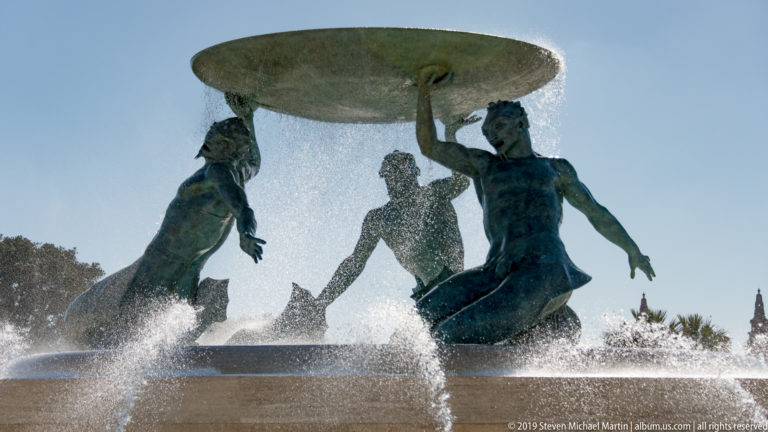 Backlit bronze figures supporting a broad fountain basin as water pours over the rim and sprays across the foreground.