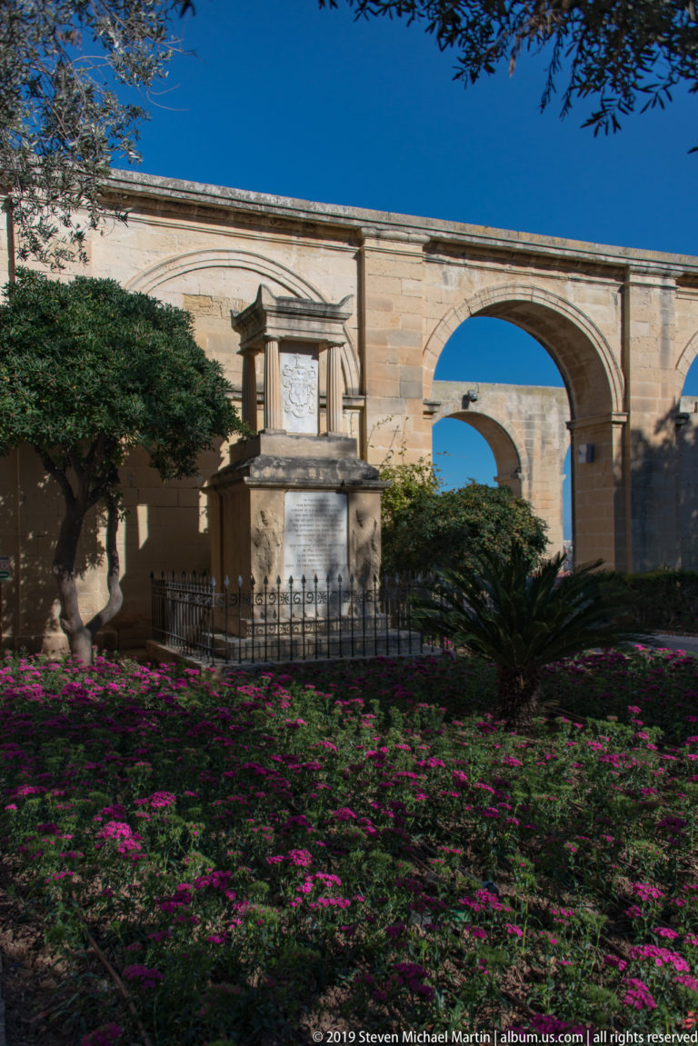 Stone monument behind iron railing in a flower-filled garden, with mature trees and two large limestone arches in the background.