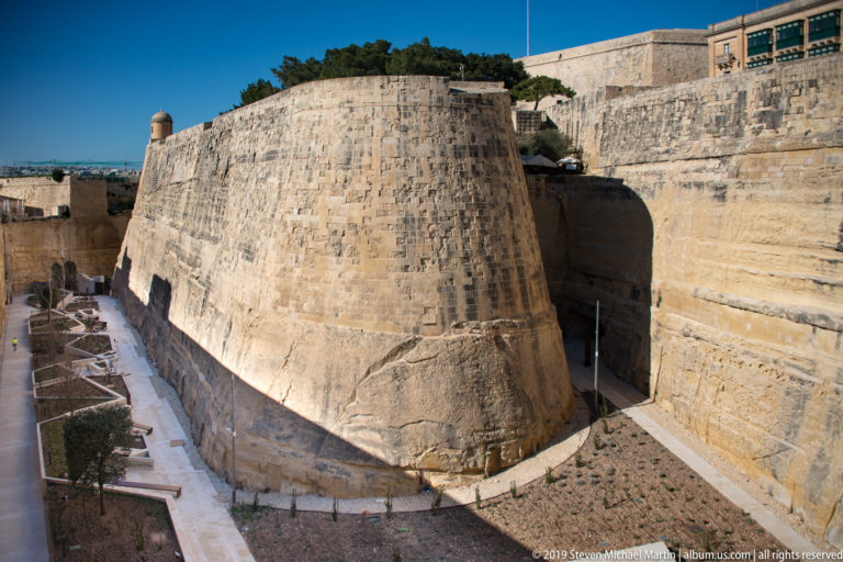 Large rounded limestone bastion rising from a dry defensive ditch, with paths, planted beds, and higher fortification walls behind.