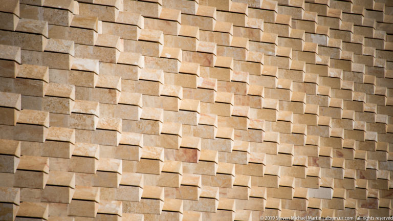 Close view of pale limestone wall formed by hundreds of rectangular blocks projecting at varied depths and casting angular shadows.