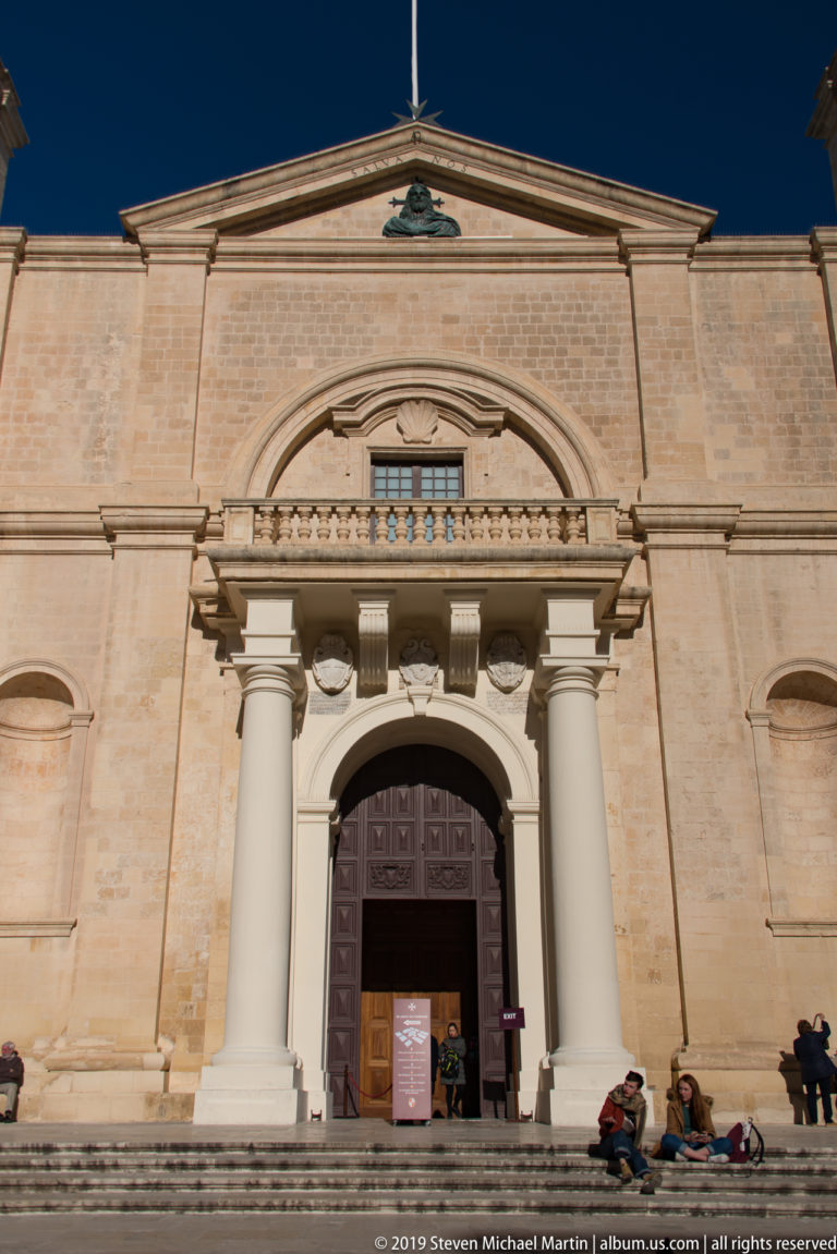 Large limestone church façade with stepped approach, arched doorway, paired columns, balcony, pediment, and visitors seated near the entrance.