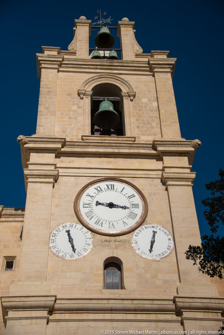 Low-angle view of a limestone clock tower with large Roman-numeral clock, two smaller dials, and open bell stages above.