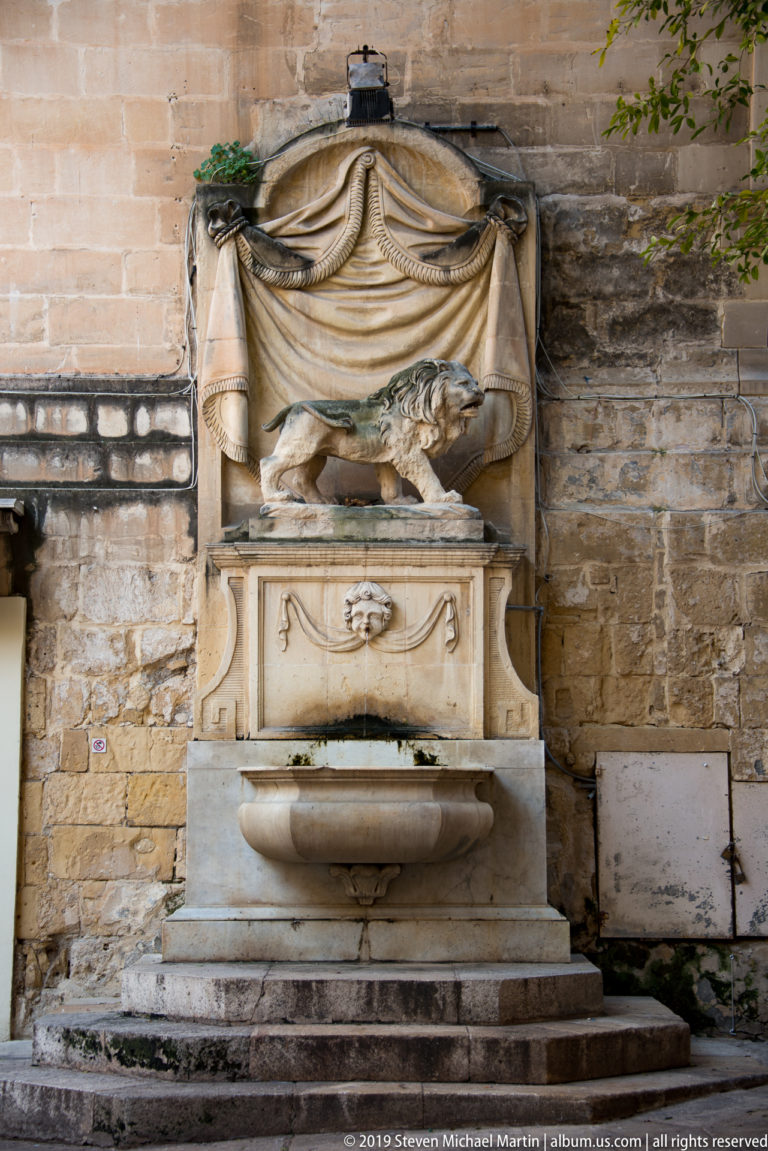 Wall fountain with reclining carved lion beneath a draped stone canopy and a shallow basin against weathered limestone masonry.