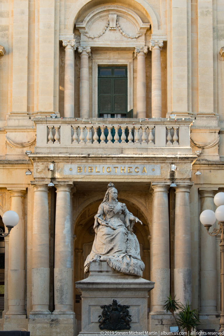 Seated stone monument before the columned entrance of the Bibliotheca, with the building name visible above the arch.
