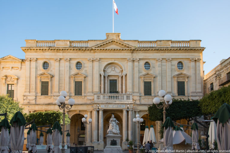 Formal limestone library façade with pediment, round windows, balcony, central monument, café umbrellas, and symmetrical classical detailing.