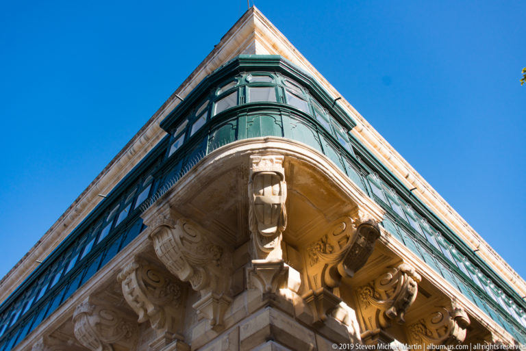 Low-angle corner view of a green enclosed timber balcony supported by carved limestone corbels and scrollwork against blue sky.