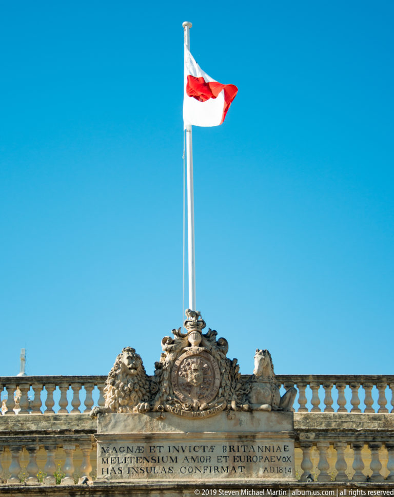 Maltese flag above a carved stone crest, animal supporters, balustrade, and Latin inscription against a clear blue sky.