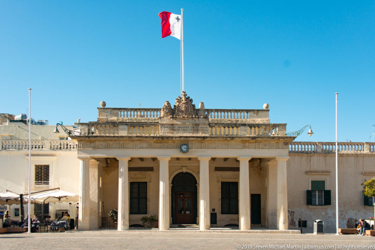 Symmetrical limestone civic building with columned portico, balustrade, sculptural crest, clock, and Maltese flag above the entrance.