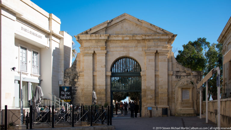 Tall limestone gateway with triangular pediment, paired pilasters, and barred arched opening leading toward people and trees beyond.