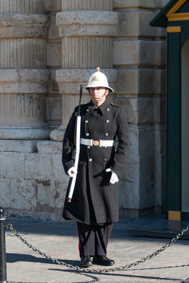 Ceremonial guard in dark uniform, white gloves, and white helmet standing with a rifle before rusticated limestone masonry.