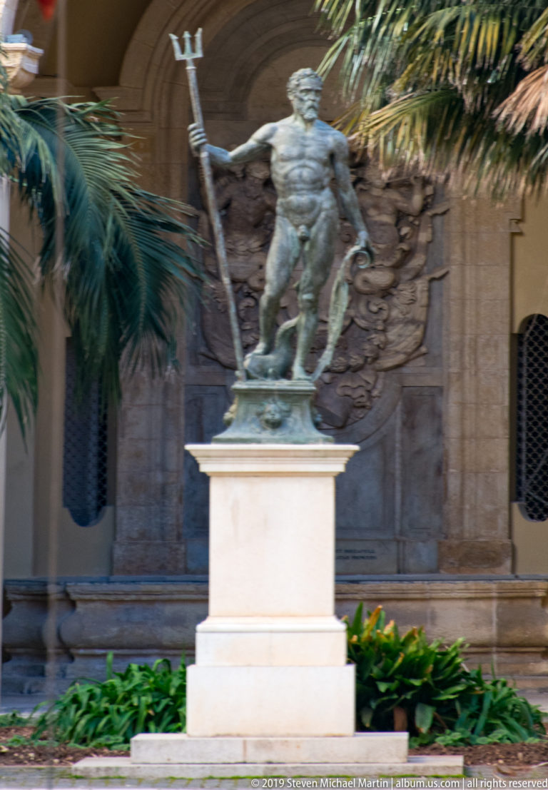 Bronze classical male figure holding a long trident-like staff beside a smaller figure, on a pale pedestal among palm trees.