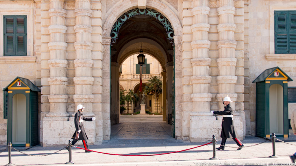 Two ceremonial guards and green sentry boxes flanking a tall rusticated limestone arch leading into a courtyard.