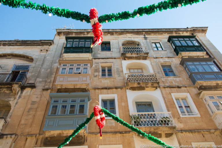 Upward view of limestone residences with projecting painted timber balconies, iron balconies, shuttered windows, and red-and-green garlands.