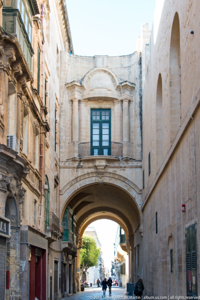 Large enclosed limestone arch spanning a narrow street, with shuttered window and balustrade above and pedestrians beyond.