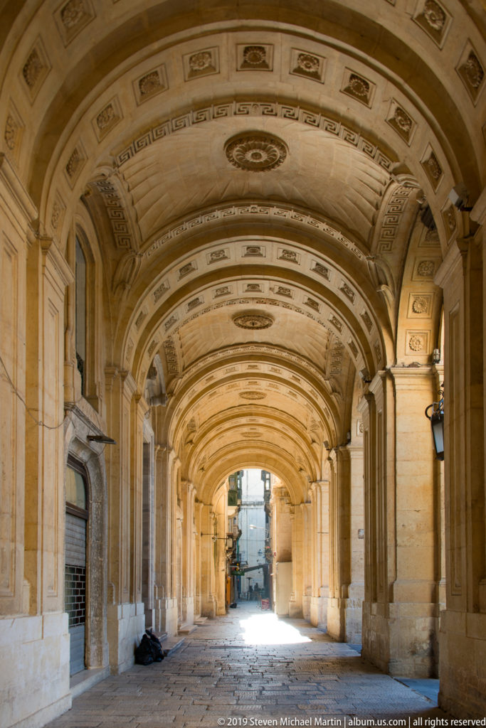 Long limestone passage with repeated arches, decorated barrel vault, stone paving, and bright opening at the far end.