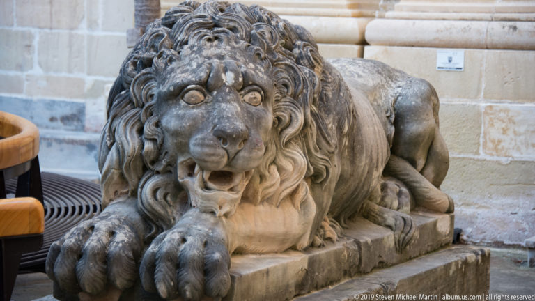 Weathered recumbent stone lion with carved mane, open mouth, and large forepaws resting on a rectangular plinth.