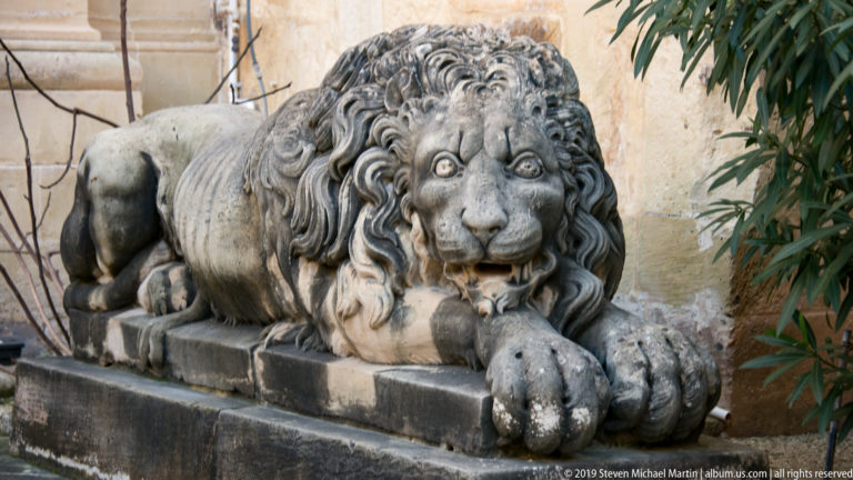 Three-quarter view of a weathered recumbent stone lion with carved mane, open mouth, and large forepaw extending forward.