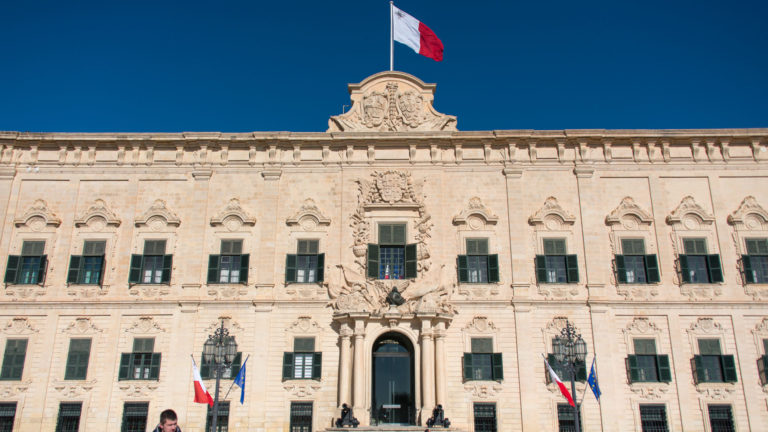 Upper façade of Auberge de Castille with green shutters, carved central ornament, heraldic roof crest, and Maltese flag against blue sky.
