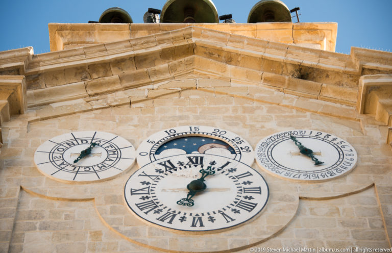Pale limestone tower with one large Roman-numeral clock and two smaller circular dials, with bells visible above the parapet.