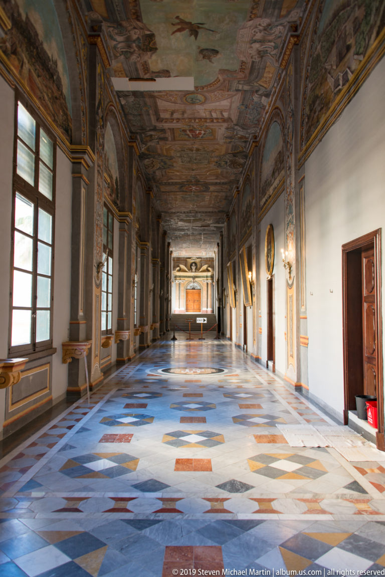 Long palace corridor with frescoed ceiling, tall windows, patterned marble floor, portraits, and a distant doorway.