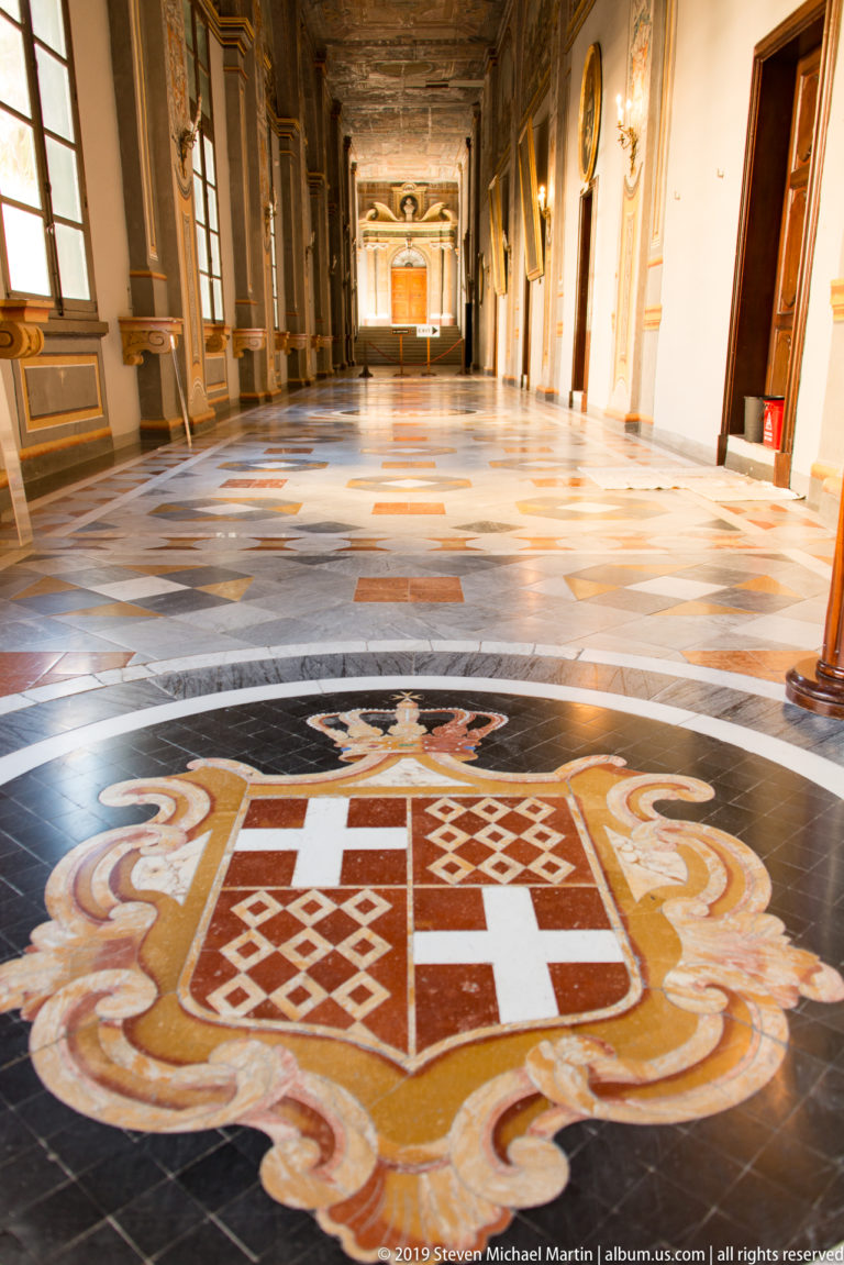 Large heraldic floor inlay in a frescoed palace corridor, with a quartered shield, scrolling border, and long hall beyond.