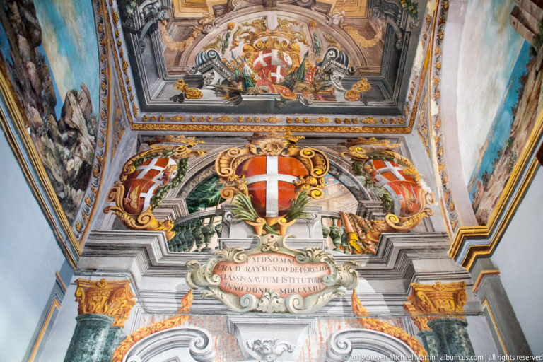 Ornate heraldic decoration above an arch with painted shields, red-and-white cross, sculpted scrollwork, inscription, and surrounding frescoes.