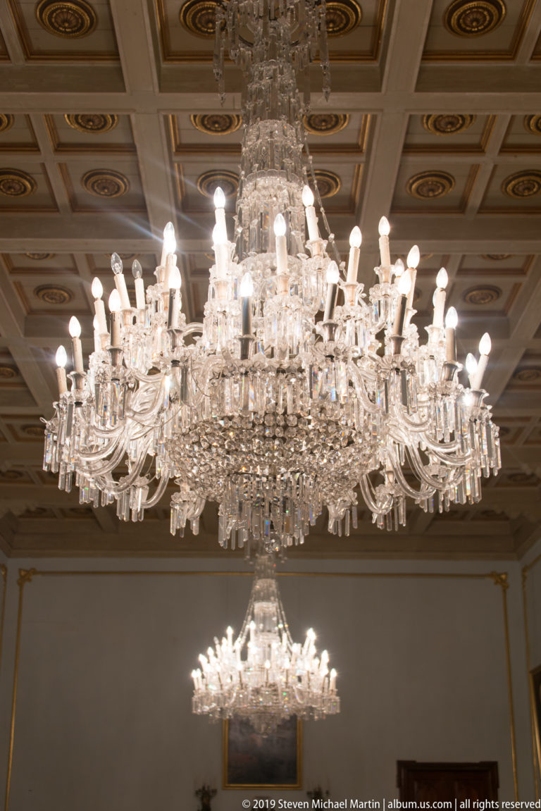 Large illuminated crystal chandelier hanging below a decorated coffered ceiling, with a second chandelier visible farther into the room.