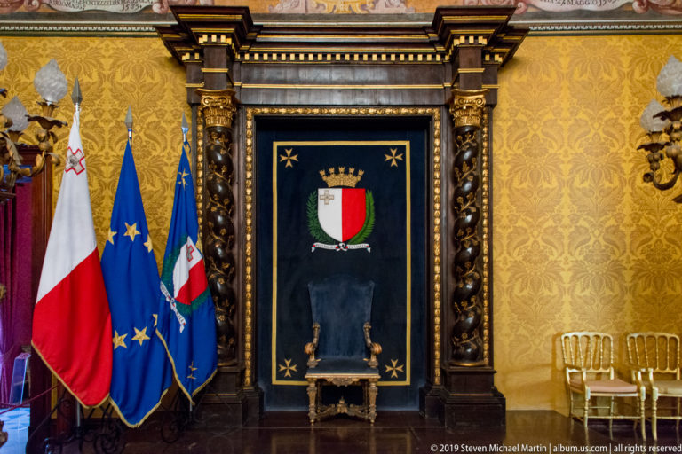 Maltese and European Union flags beside a dark ceremonial chair beneath a crowned Maltese shield in an ornate palace interior.