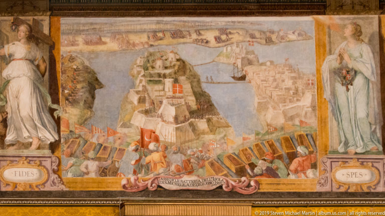 Large palace wall painting of a coastal siege with ships, fortifications, soldiers, banners, and allegorical female figures at both sides.