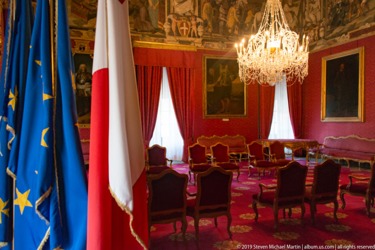 Formal red palace room with crystal chandelier, gilded chairs, red curtains, portraits, and Maltese and European Union flags.