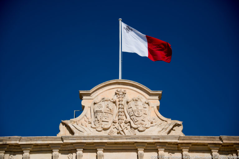 Maltese flag flying above an ornate carved heraldic crest and shield at the roofline of the Auberge de Castille.