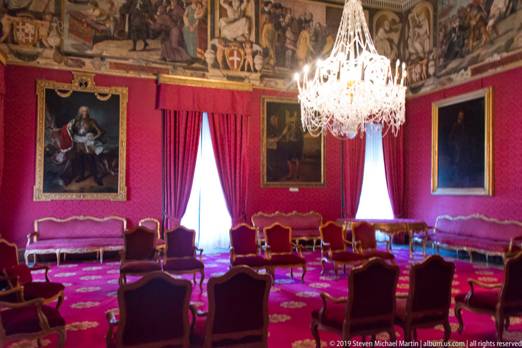 Crimson ceremonial room with chandelier, gilt-framed portraits, upholstered chairs and settees, and painted decoration along the upper walls.