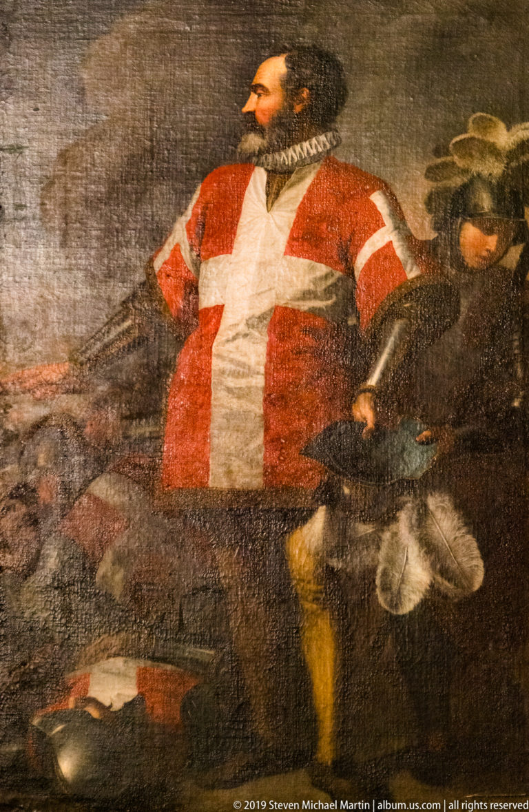 Painted bearded man wearing a red surcoat with a large white cross and ruff collar, surrounded by partially visible figures.