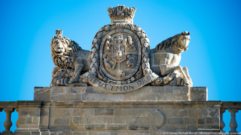 Stone royal coat of arms above a balustrade, with crowned shield, lion and horse supporters, and motto ribbon against blue sky.