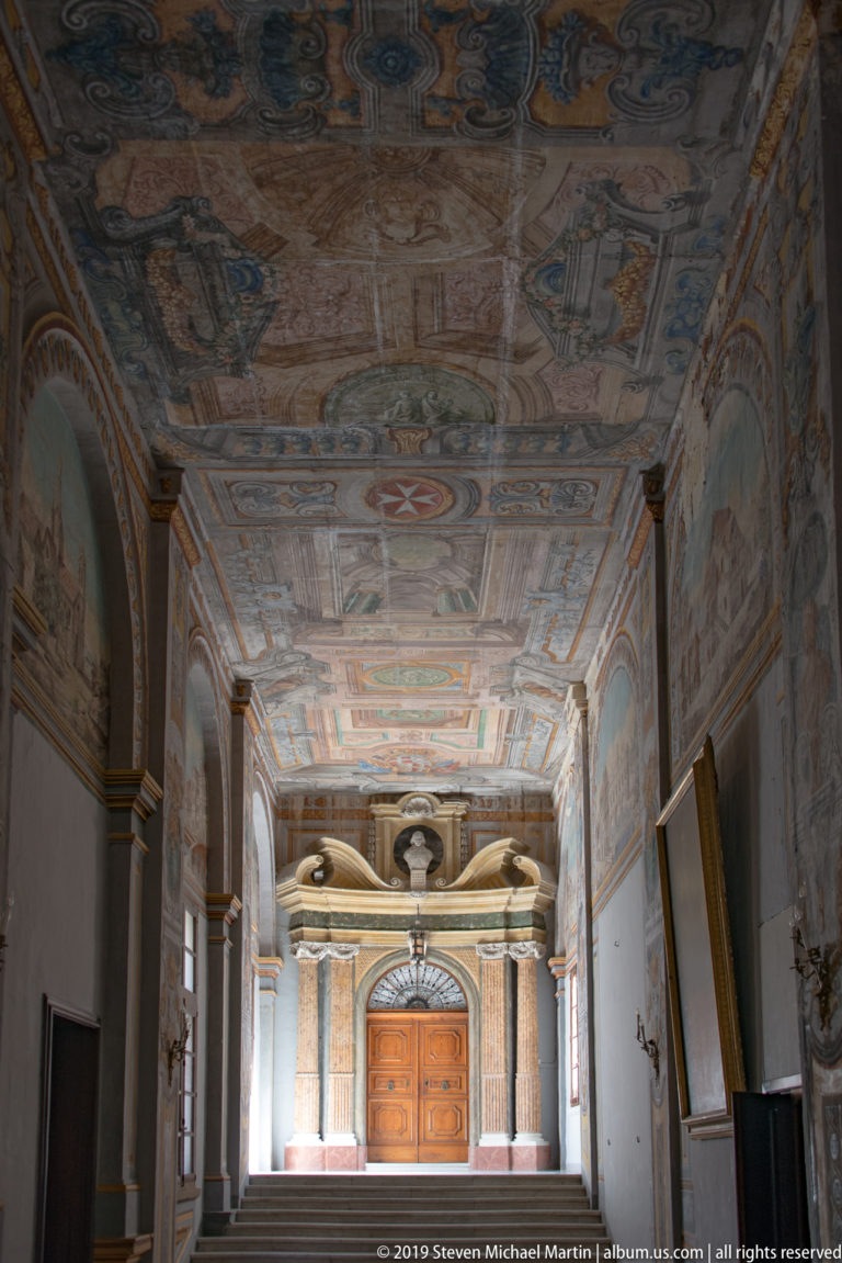 Long palace corridor with painted ceiling panels, pilasters, arches, busts, framed works, and a monumental doorway at the far end.
