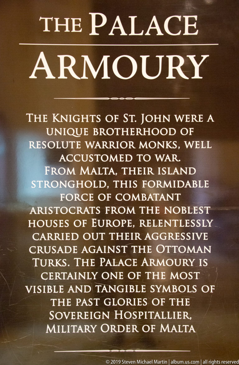 Dark museum panel titled “The Palace Armoury,” with centered white text describing the Knights of St. John and their arms collection.