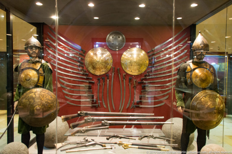 Two armored figures flank a red museum display filled with swords, round shields, helmets, firearms, and other historic weapons.