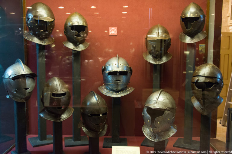 Red-backed display case containing numerous historic metal helmets with varied brims, visors, face openings, and neck protection.