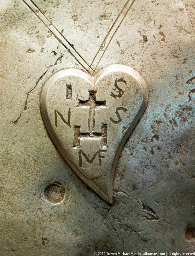 Heart-shaped marker set into a worn pale surface, carved with a cross, stepped symbol, and letters around its edges.